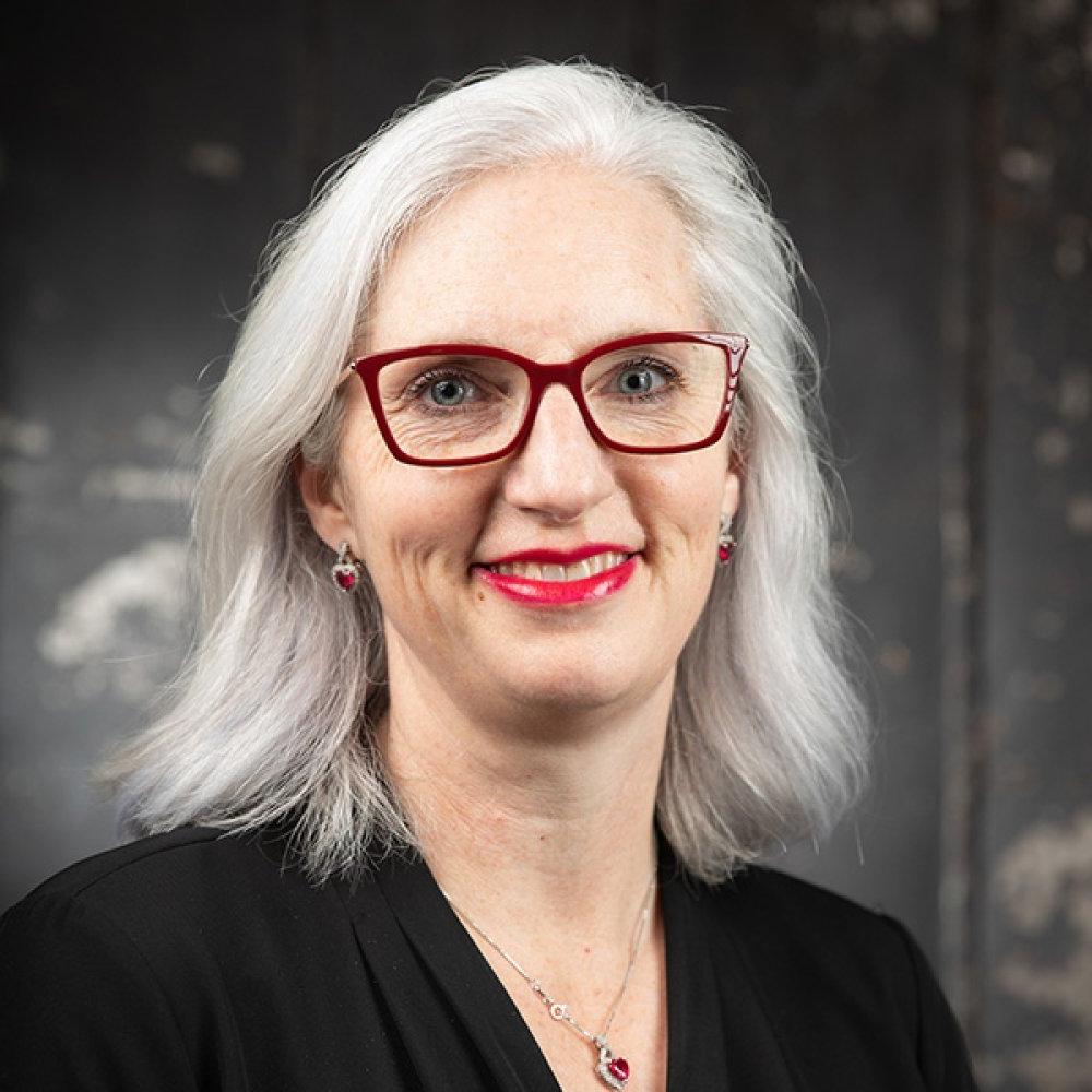 Lady with shoulder length silver hair wearing red glasses, necklace, black top in front of black studio background